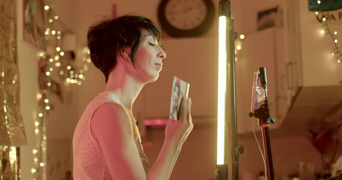 A woman uses the front camera of a mobile phone. She Shows photos online to the interlocutor. Strip light lamp. Do a halfcast in the evening.