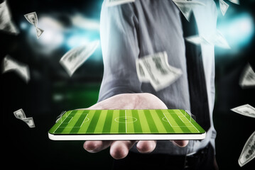 Businessman, with banknotes around, holds cellphone in his hand with a football field on the display