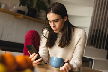 Non-binary trans woman holding a coffee cup using smartphone in the kitchen at home