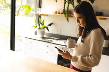 Non-binary trans woman holding a coffee cup using smartphone in the kitchen at home
