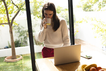 Non-binary trans woman drinking coffee and using laptop in the kitchen at home