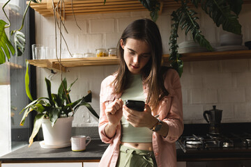 Thoughtful non-binary trans woman using smartphone in the kitchen at home