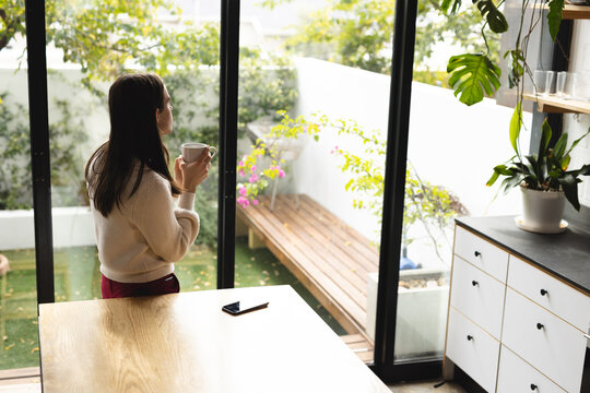Thoughtful non-binary trans woman holding a coffee cup looking out of the window at home