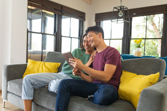 Diverse gay male couple smiling while using digital tablet sitting together on the couch at home