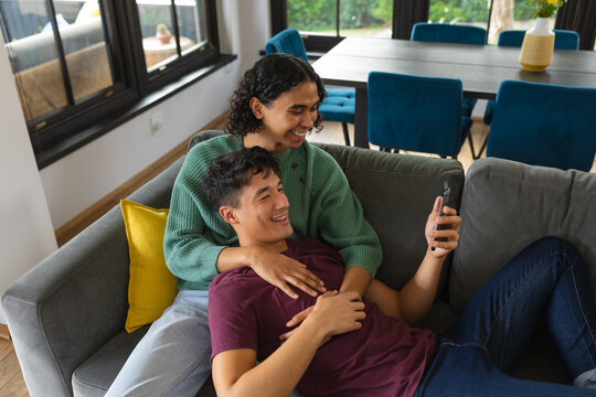 Diverse gay male couple smiling while using smartphone sitting together on the couch at home