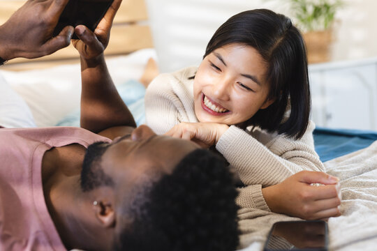 Happy diverse couple lying in bed and using smartphones