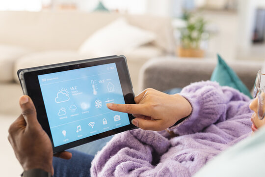 Happy diverse couple sitting on sofa in living room and using tablet with smart home interface