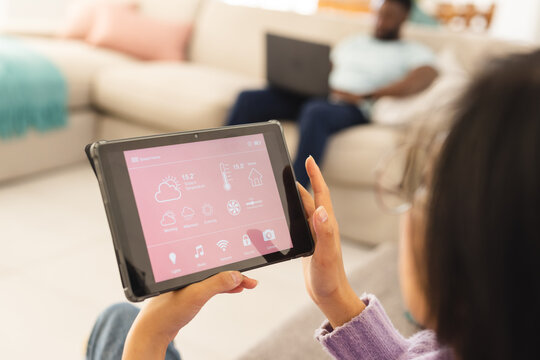 Asian woman sitting on sofa in living room and using tablet with smart home interface