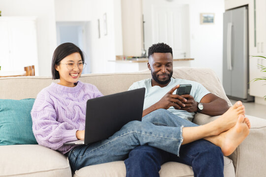 Happy diverse couple sitting on sofa in living room and using laptop and smartphone