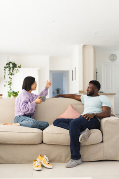 Happy diverse couple sitting on sofa in living room, drinking coffee and talking