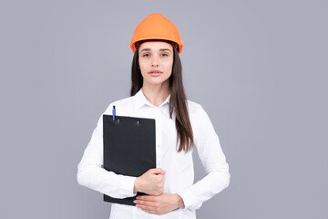 Serious woman worker in protective helmet with clipboard isolated on grey background. Young woman construction manager. Architect woman, female worker in hardhat helmet.