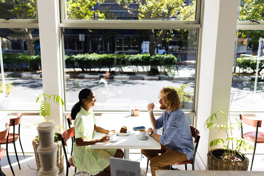 Happy diverse couple sitting at table and talking with cup of coffee in cafe