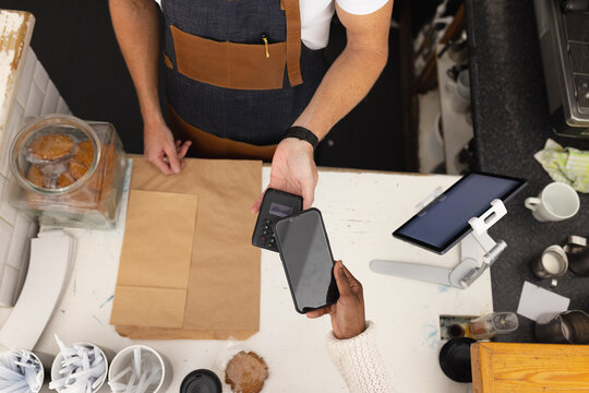 Diverse male barista and woman paying with smartphone in cafe