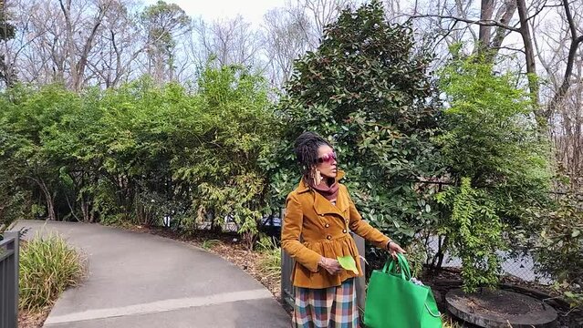 Slow motion footage of an African American woman with long sisterlocks wearing a brown coat and sunglasses walking in the garden with trees and plants at Atlanta Botanical Garden in Atlanta Georgia