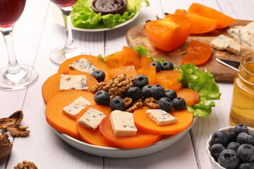 Delicious persimmon with blue cheese, blueberries, lettuce and nuts on white wooden table, closeup