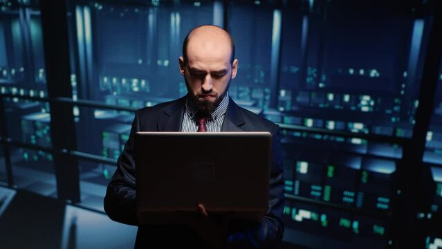 System engineer running hosting server diagnostics with laptop, standing in modern big data center space. IT technician working on professional mainframe hardware. Tripod shot.