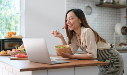 Pretty asian woman eating vegetable salad  and using laptop at table kitchen. Healthy nutrition...