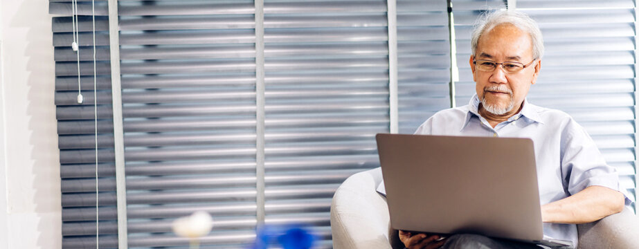 Senior Asian Man Having Good Time Work Using Laptop Computer.Happy Elderly Man Checking Social Media And Reading News Or Shopping Online While Sitting On Sofa At Home