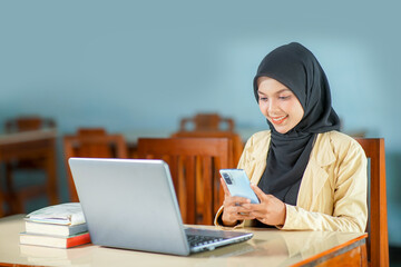beautiful young asian woman wearing hijab smiling at work, operating a mobile phone and laptop computer at her desk.