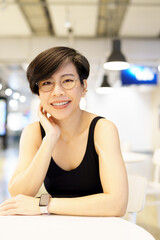 Stylish, happy and confident short hairstyle Asian woman wear eyeglasses, blank casual tank top smiling, sitting, looking at camera in beautiful, modern cafe for health, wellbeing, self care concept