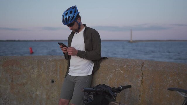 Theme is cycling in seaside. Male cyclist stopped to enjoy the sea view and see the road on the map on his smartphone. A male with a bicycle on the embankment is resting and using a smart phone.