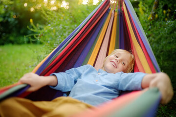 Cute little blond white boy enjoy and having fun with multicolored hammock in backyard or outdoor playground. Summer outdoors active leisure for kids. Child relaxing and swinging in hammock.