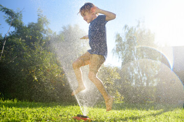 Funny little boy playing with garden sprinkler in sunny backyard. Preschooler child laughing, jumping and having fun with spray of water. Summer holidays for kids in the village - obrazy, fototapety, plakaty