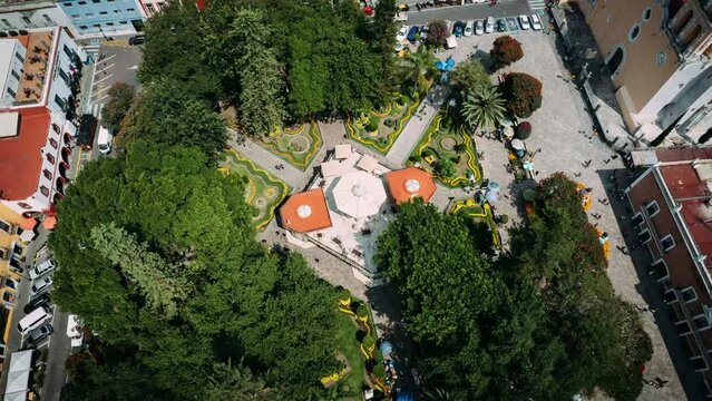 Drone moving around the kiosk of mexican town Atlixco surround by trees and people passing one evening