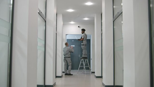 Two multi ethnic installers in uniform sets up security camera in office hallway using laptop. They talk, check CCTV cameras in computer program. Concept of surveillance system and privacy. Dolly shot