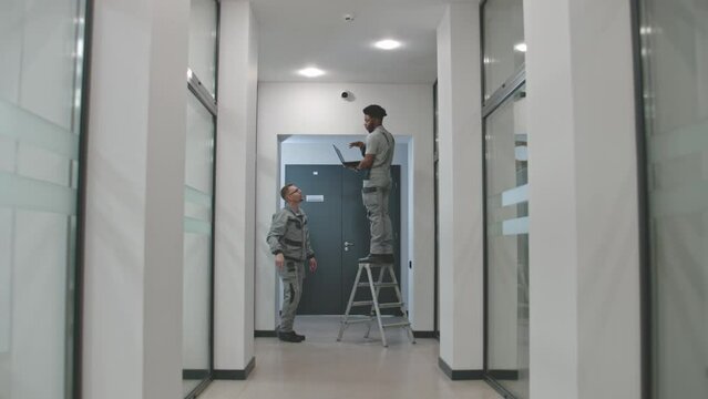 Two multi ethnic installers in uniform sets up security cameras in office corridor using laptop. They talk, check CCTV cameras in computer program. Concept of monitoring system and privacy. Dolly shot