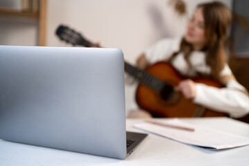 Music college female student practicing acoustic guitar exercise, reading notes from laptop...