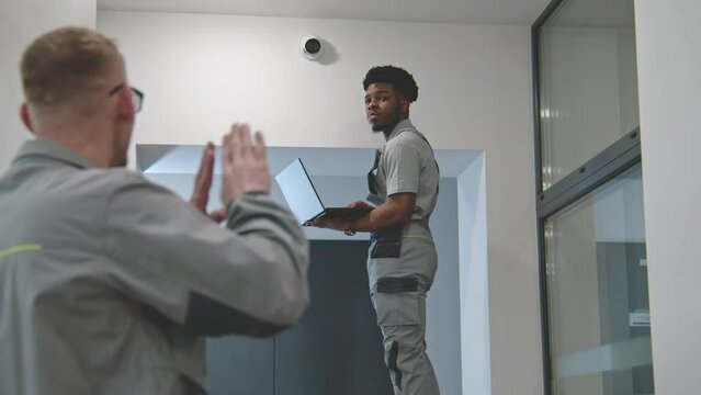 Two multi ethnic installers in uniform sets up security camera in office hallway using laptop. They talk, check CCTV cameras in computer program. Concept of surveillance system and privacy. Dolly shot