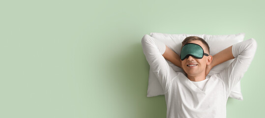Happy young man with sleep mask and pillow on green background with space for text