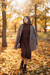 Happy trendy woman in gray coat outdoors in the city park in autumn. Attractive free girl in park with foliage in background.