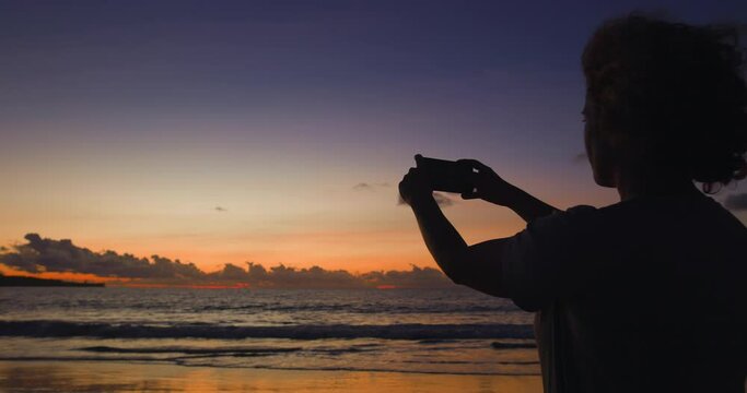 Curly-haired woman with phone in her hands takes pictures of beautiful summer sunset. Ocean waves reflect rays of golden setting sun, beautiful picture of sunset on ocean. Woman takes photo on phone