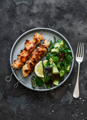 Chicken bacon skewers and  avocado, arugula, spinach, chard salad on a dark background top view