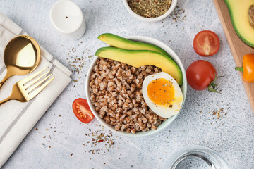 Bowl of tasty buckwheat porridge with soft boiled egg and fresh vegetables on grey table