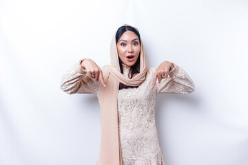 Shocked Asian Muslim woman wearing hijab pointing at the copy space below her, isolated by white...