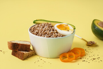 Bowl of tasty buckwheat porridge with soft boiled egg, fresh vegetables and bread on yellow background