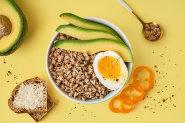 Bowl of tasty buckwheat porridge with soft boiled egg, fresh vegetables and bread on yellow background