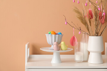 Vase with tree branches, Easter eggs and pampas grass on table near beige wall