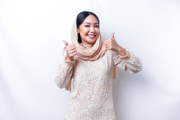 Excited Asian Muslim woman wearing a hijab gives thumbs up hand gesture of approval, isolated by...