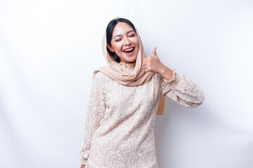Excited Asian Muslim woman wearing a hijab gives thumbs up hand gesture of approval, isolated by...