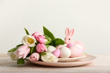 Plate with Easter eggs, tulip flowers and bunny on table near light wall