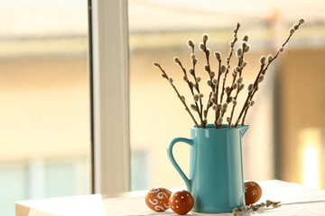 Jug with pussy willow branches and Easter eggs on table near window