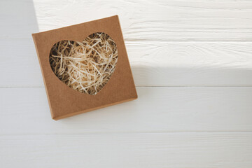 Eco gift wrapping. Cardboard brown gift box with a transparent heart and cut paper inside on white wooden background. Flat lay