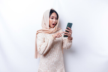 Shocked Asian woman wearing hijab, holding her phone, isolated by white background