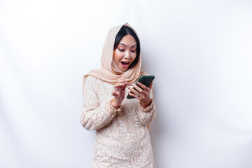 Shocked Asian woman wearing hijab, holding her phone, isolated by white background