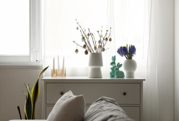 Vases with tree branches, Easter eggs, flowers and rabbits on chest of drawers in room