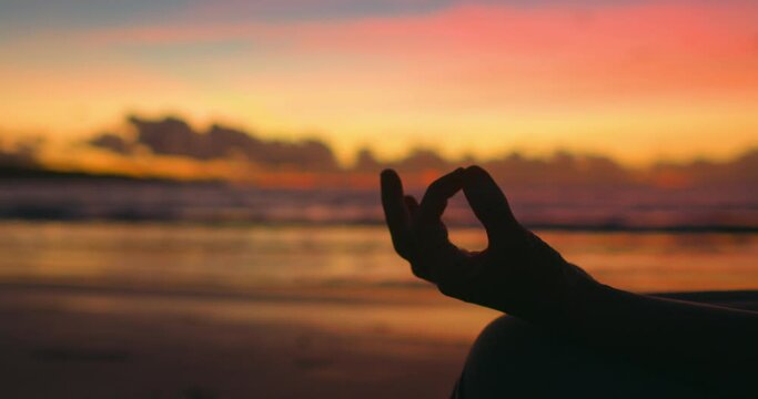 Silhouette of woman's hands, sea surf, sunset in pink, orange, red tones. Lotus pose, main asana in yoga, thumb and index finger are joined. Gyan mudra. Woman does yoga on beach in setting sun